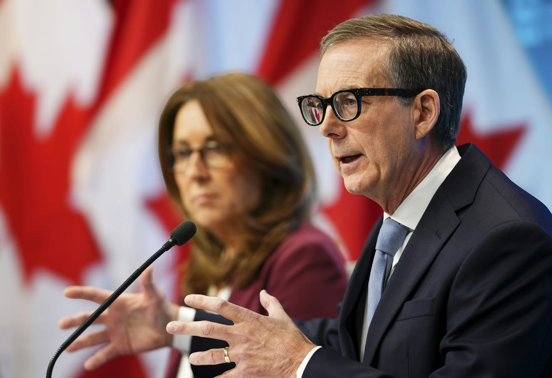 An older man in a suit speaks into a microphone while a woman sits beside him. Behind him, Canadian flags can be seen.