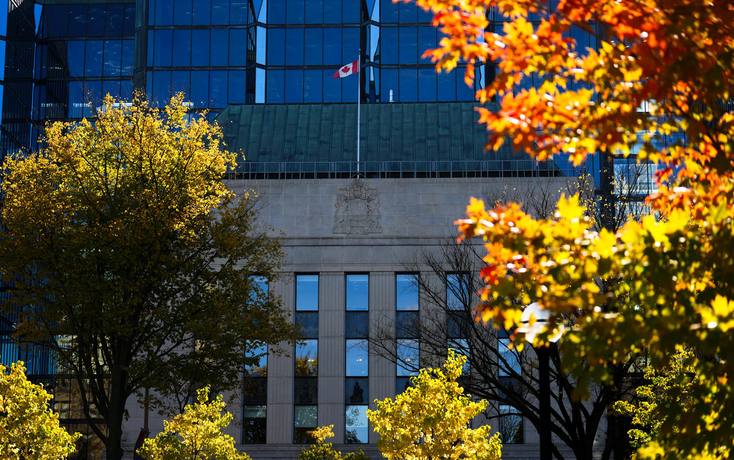 A building with a Canadian flag on the front is framed by deciduous trees with green leaves that are starting to turn orange and red
