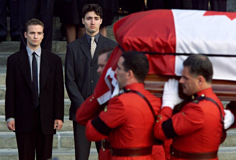 Two young men, one weeping, watch as a flag-draped coffin is walked to a hearse by Mounties in red serge jackets.