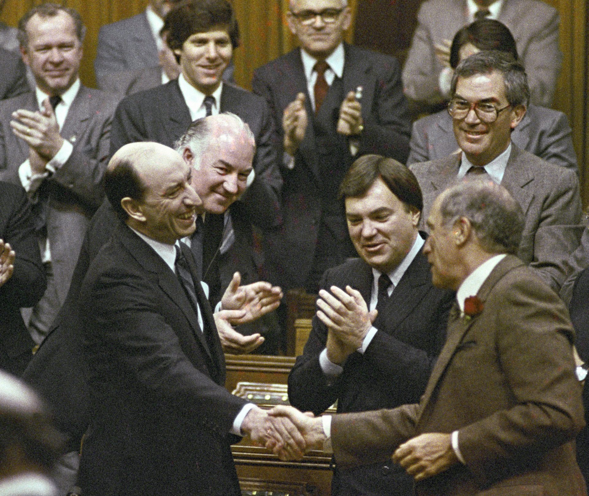 A smiling bald man in a dark suit shakes the hand of another man in a brown suit jacket in the House of Commons.