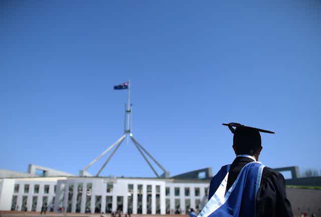 A student in graduation robes  stands in front of Parliament House in Canberra. 