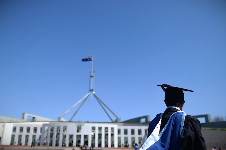 A student in graduation robes stands in front of Parliament House in Canberra.
