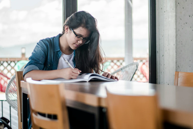 A young woman sits at a table, writing in a book.