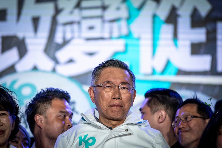 A close-up shot of a Taiwanese man wearing glasses and dressed in a white jacket at a rally.