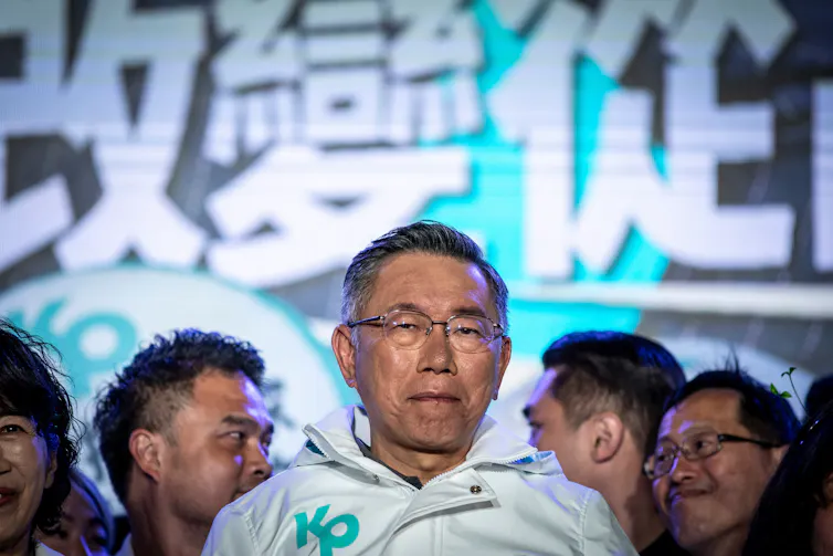 A close-up shot of a Taiwanese man wearing glasses and dressed in a white jacket at a rally.