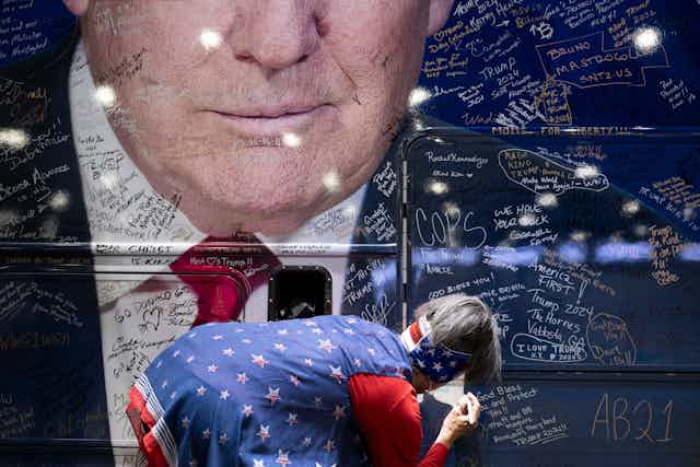 A person wearing a blue shirt with red and white stars and an American flag bandana bends down to write something on a large blue bus, which shows the bottom half of Donald Trump's face