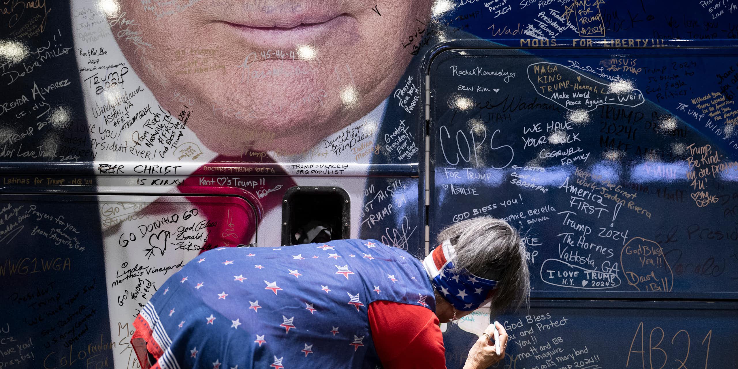 A person wearing a blue shirt with red and white stars and an American flag bandana bends down to write something on a large blue bus, which shows the bottom half of Donald Trump's face