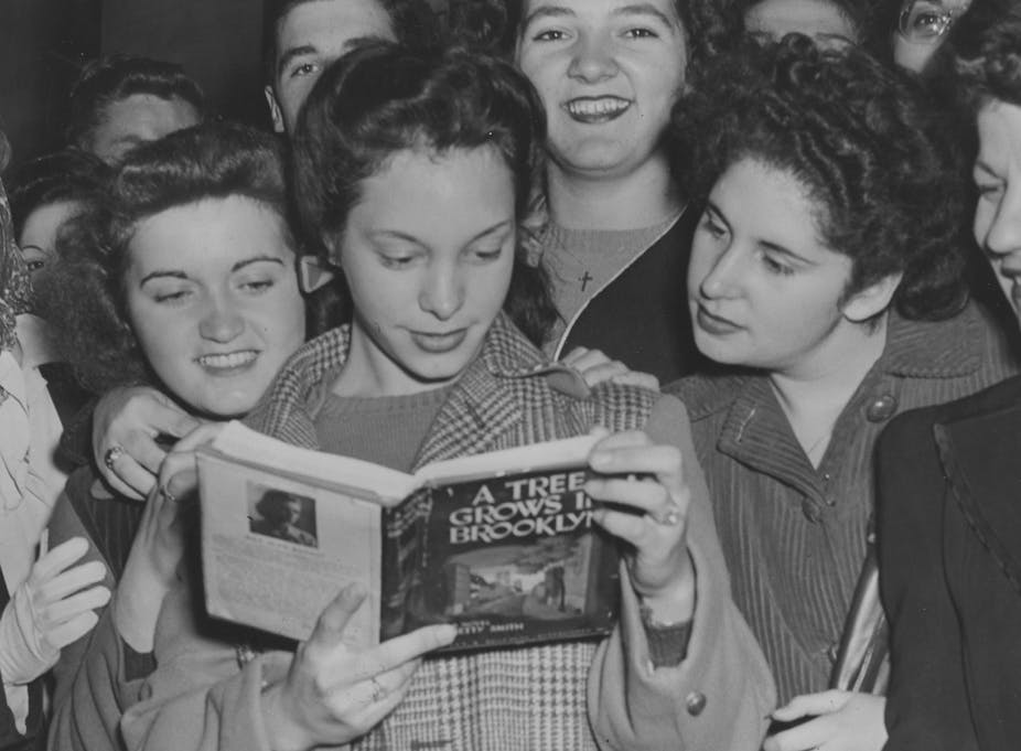 Black and white photo of a group of teen girls huddling around another girl who's reading 'A Tree Grows in Brooklyn.'