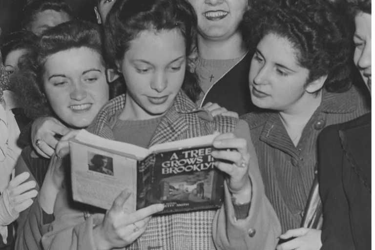 Black and white photo of a group of teen girls huddling around another girl who's reading 'A Tree Grows in Brooklyn.'