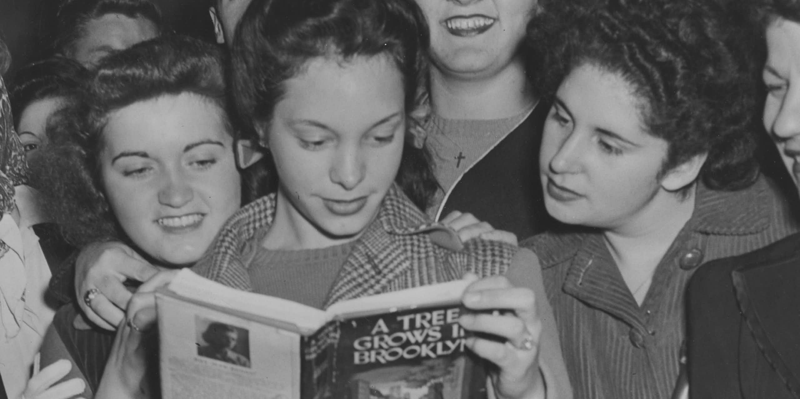 Black and white photo of a group of teen girls huddling around another girl who's reading 'A Tree Grows in Brooklyn.'