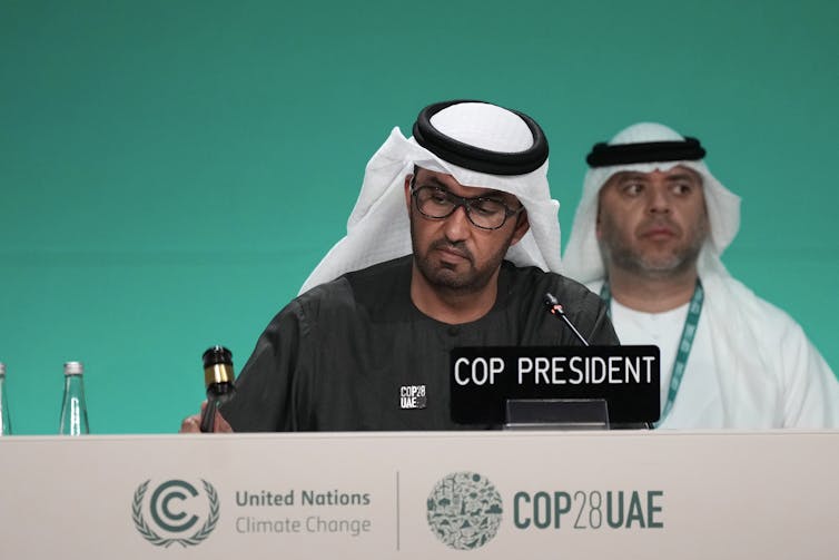 A south Asian man wearing glasses in a white kufiyah bangs a small gavel on a table with the COP logo printed on the front. A name plate in front of him says COP PRESIDENT.