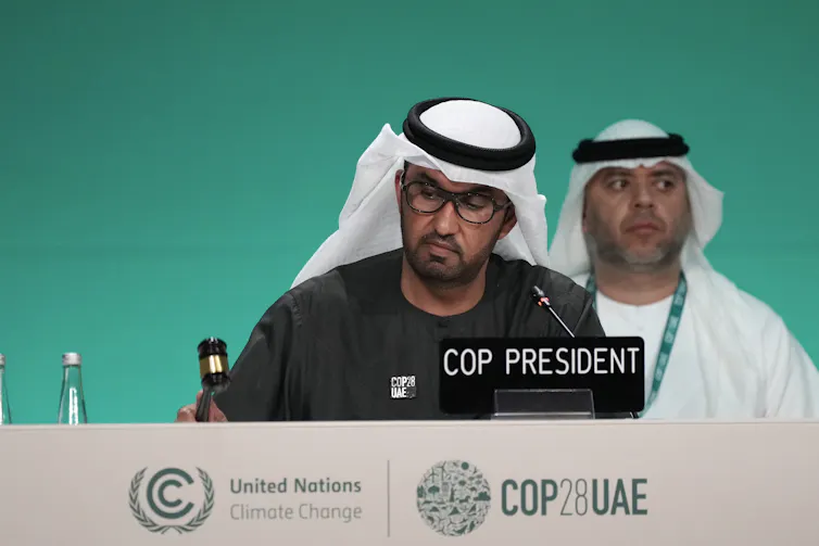 A south Asian man wearing glasses in a white kufiyah bangs a small gavel on a table with the COP logo printed on the front. A name plate in front of him says COP PRESIDENT.