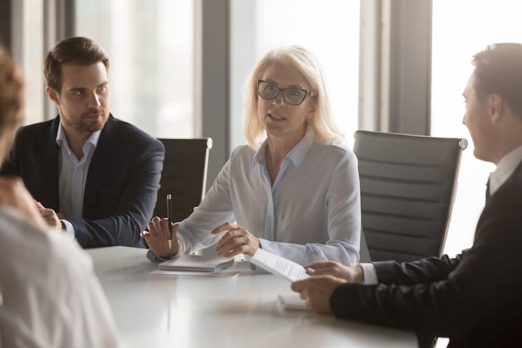 A middle aged businesswoman talks to a group of people at a conference table