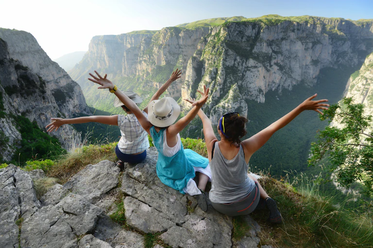 Three young women hold their arms up in the air while sitting on top of a tall mountain overlooking a valley.
