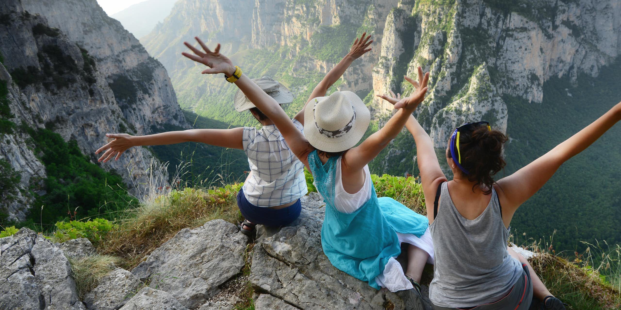 Three young women hold their arms up in the air while sitting on top of a tall mountain overlooking a valley.