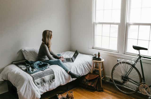 A young woman sits on her bed looking at a laptop. There is a bike nearby propped up agains the wall.