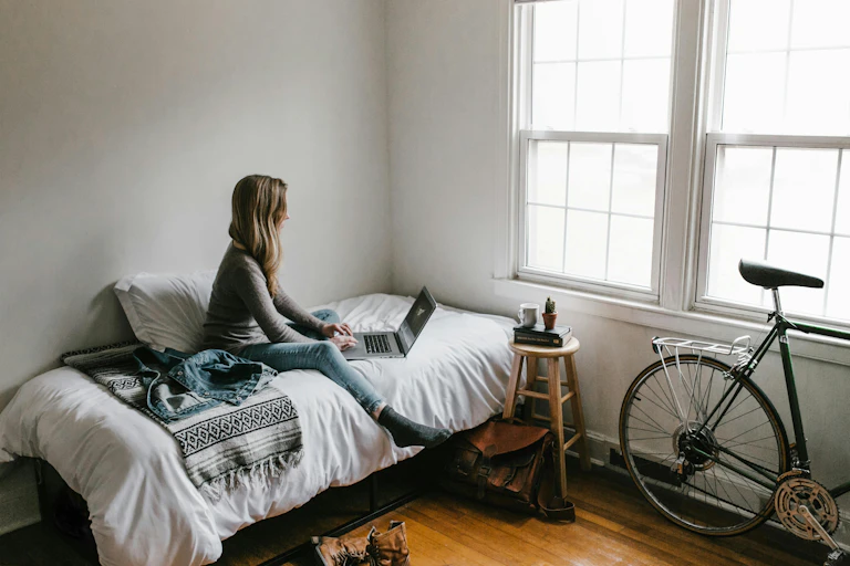 A young woman sits on her bed looking at a laptop. There is a bike nearby propped up agains the wall.