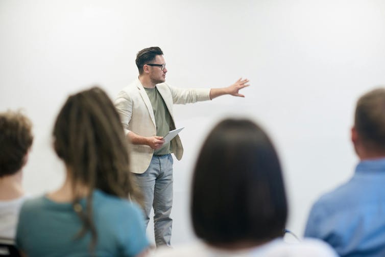 A man stands in front of a seated audience and points at a board