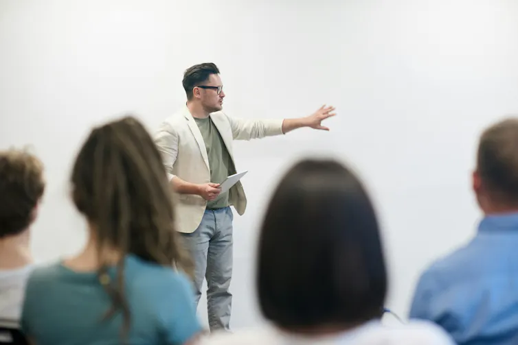 A man stands in front of a seated audience and points at a board