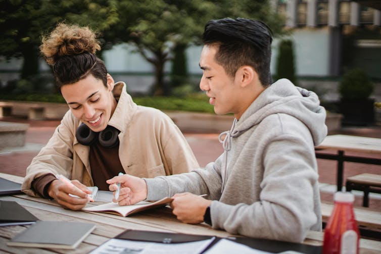 Two young men sit side by side at an outdoor table, looking at a notebook.
