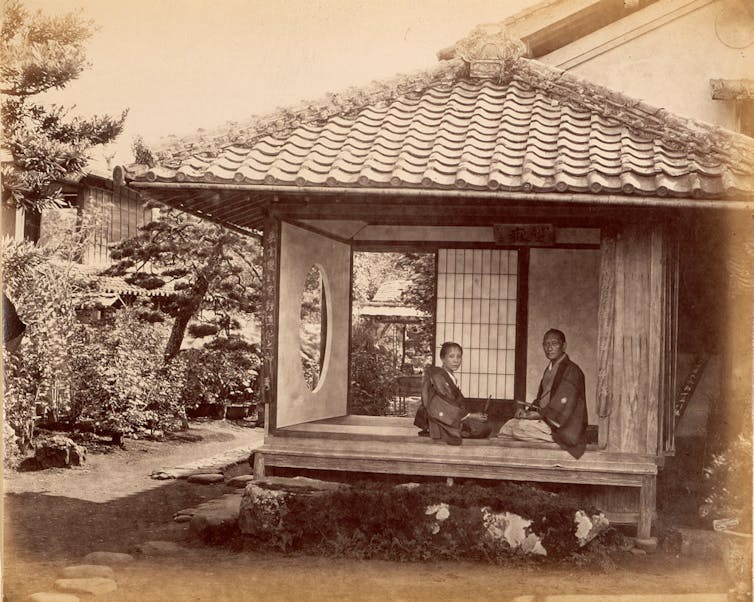 Two people kneeling in a small, roofed room open to the outdoors, set in a garden, look at the photographer.