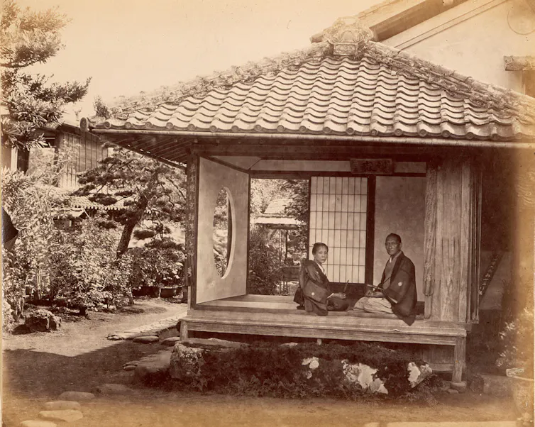 Two people kneeling in a small, roofed room open to the outdoors, set in a garden, look at the photographer.
