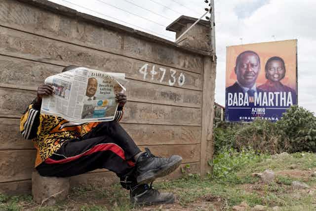 A person sits up against the wall of a house, their legs crossed, reading a newspaper. In the background there's an election billboard for two Kenyan political candidates