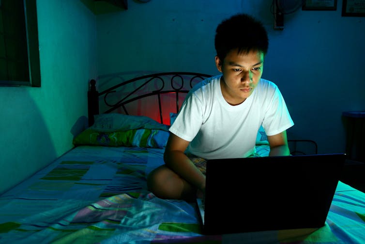 A young man uses his laptop at night while sitting on his bed.