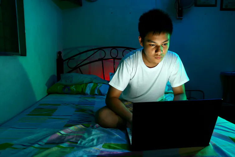 A young man uses his laptop at night while sitting on his bed.