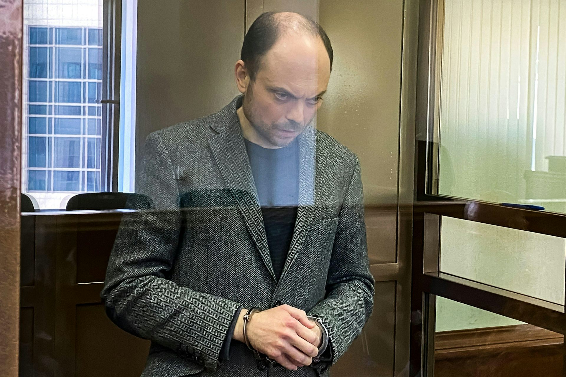 A balding man in a grey jacket stands with his hands clasped and his head down