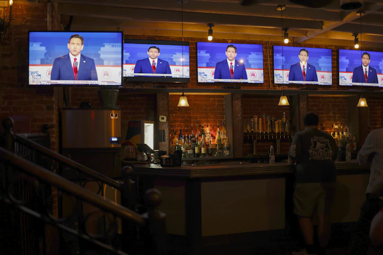 Five tv screens suspended over a dark bar, with a man shown talking in them.