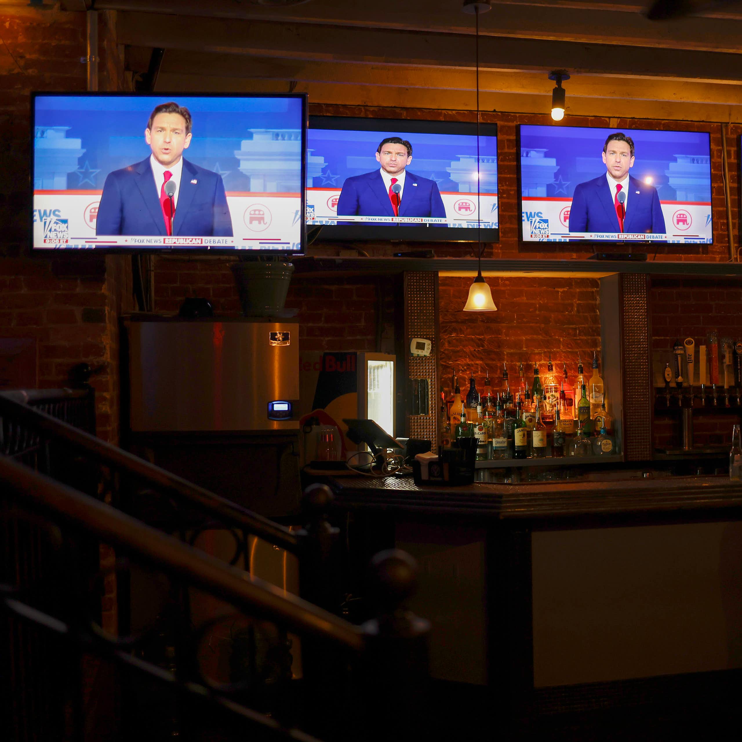 Five tv screens suspended over a dark bar, with a man shown talking in them.
