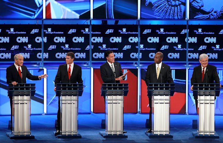 Five men debating each other on a stage behind lecterns.