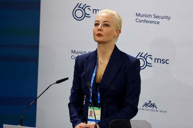 A blond woman wears a formal blue blazer and looks straight ahead, standing in front of a white backdrop with the words 'Munich Security Conferece' in blue 
