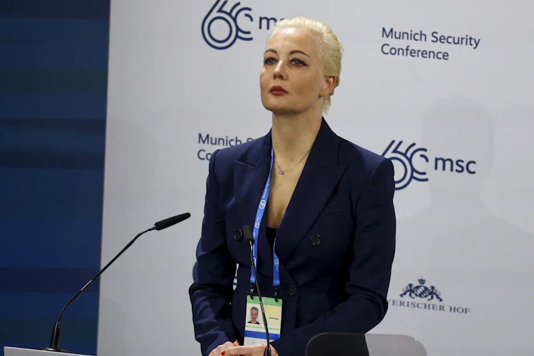 A blond woman wears a formal blue blazer and looks straight ahead, standing in front of a white backdrop with the words 'Munich Security Conferece' in blue