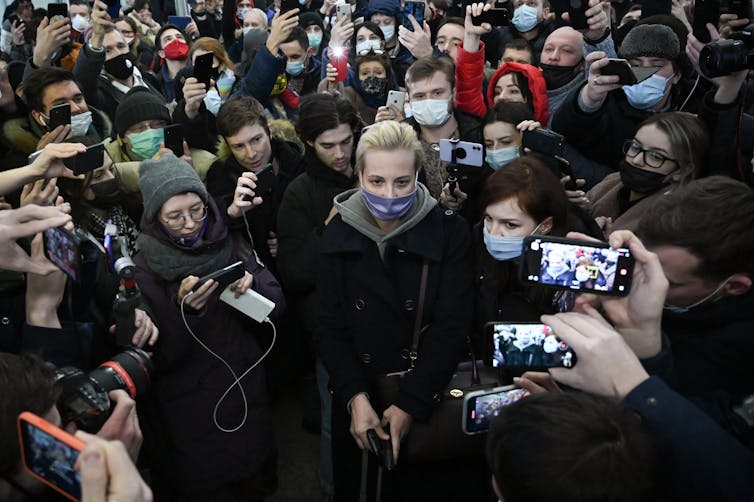 A white, blonde haired woman wears a face mask and stands in a crowd of people, who all direct cell phone cameras at her.