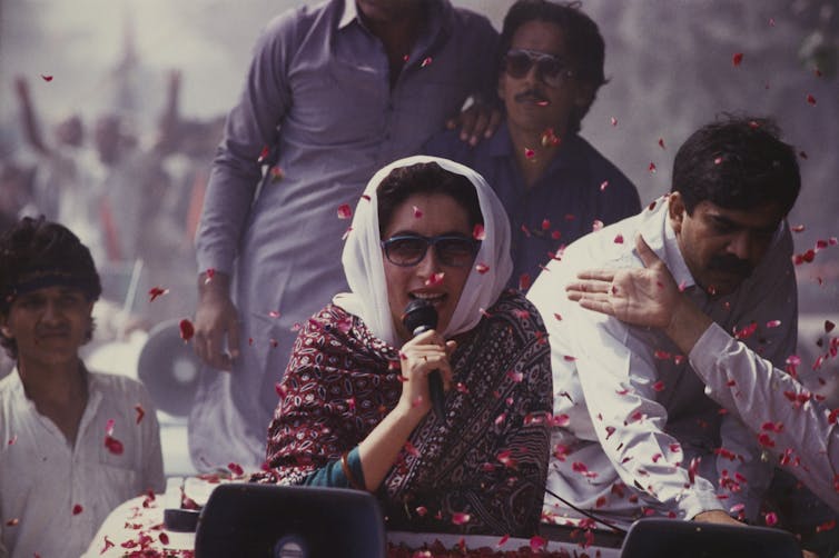 A woman in dark sunglasses and a white head cover leans against the top of a car that she rides on as she speaks into a microphone. Several men stand or sit around her.