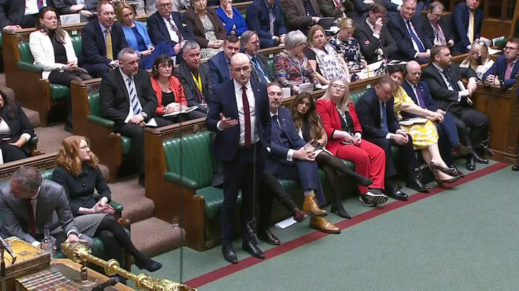 Stephen Flynn standing up on the floor of the House of Commons with SNP MPs sitting behind him on green benches.