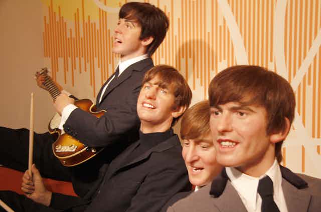 four young men in suits smiling stand in front of a pale orange background. one holds a guitar.
