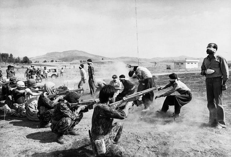 A group of men with rifles kneel in front of several men who are wearing blindfolds.