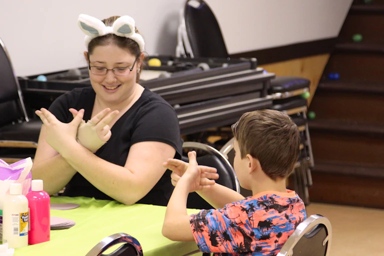 Adult seen in bunny ears at a table making a sign to a child making the same sign.