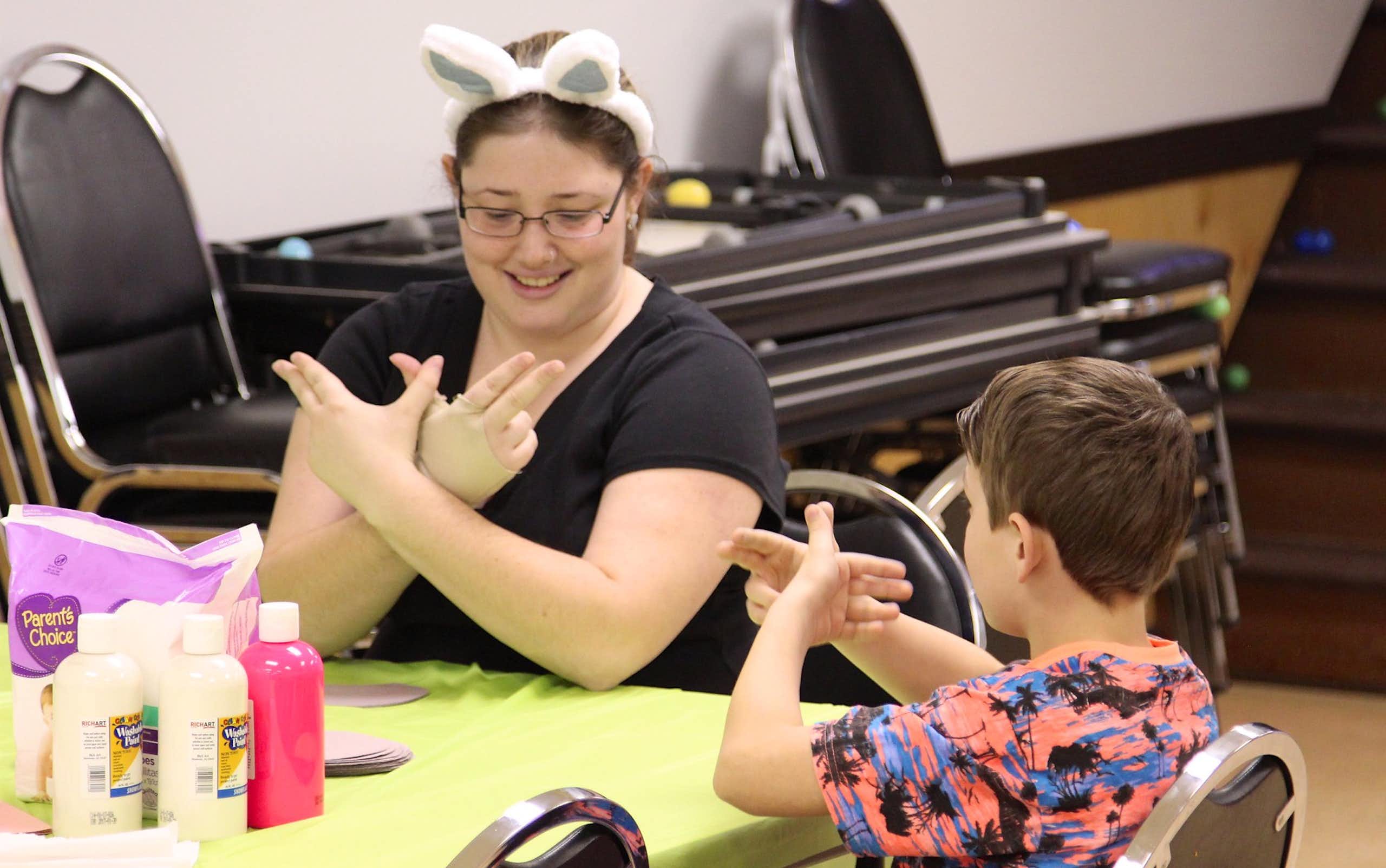 Adult seen in bunny ears at a table making a sign to a child making the same sign.