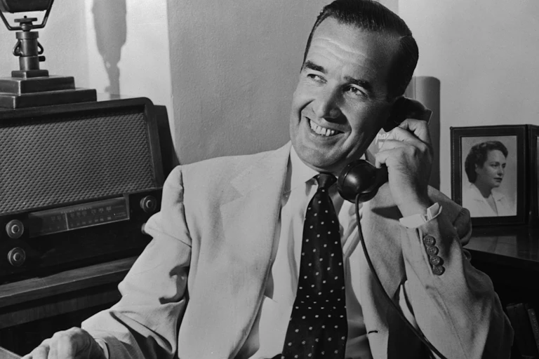 A smiling middle aged man is dressed in a lightly colored suit and is smiling as he talks on a telephone.