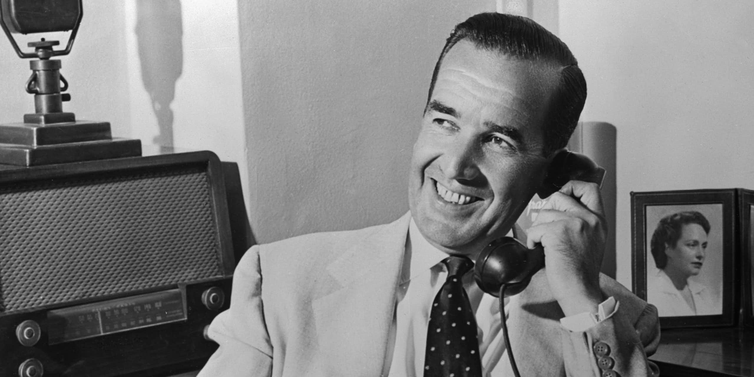 A smiling middle aged man is dressed in a lightly colored suit and is smiling as he talks on a telephone. 