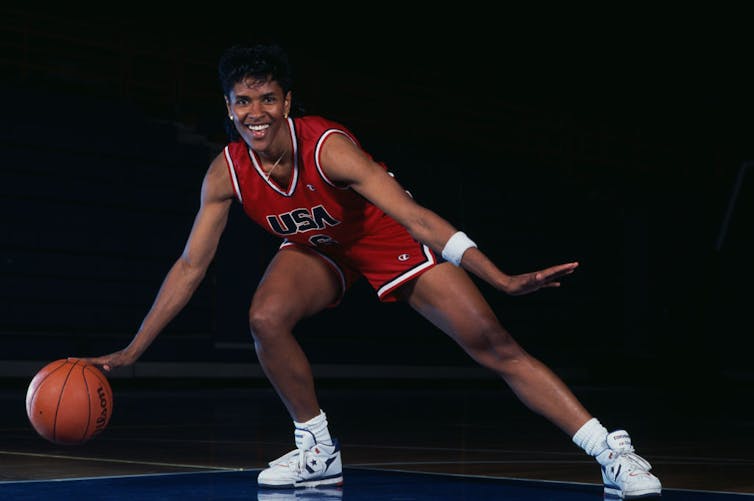 Young woman with short hair poses while dribbling a basketball and wearing a red, white and blue Team USA jersey.
