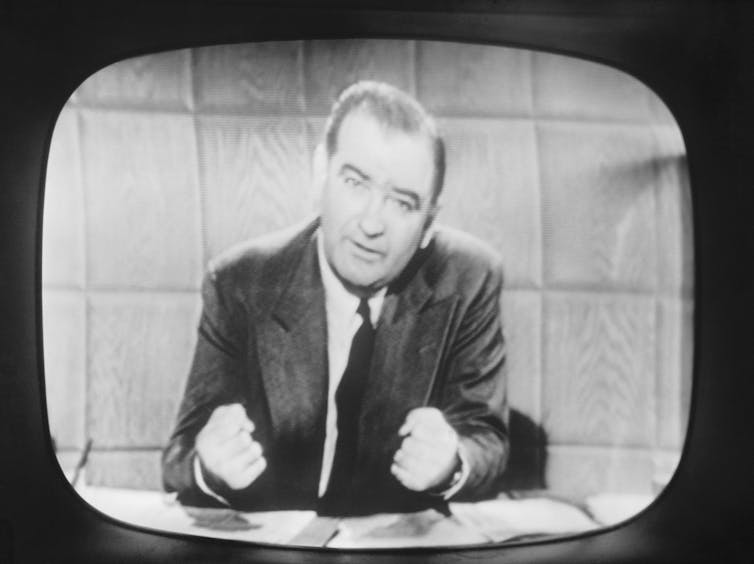 Un hombre blanco se sienta detrás de un escritorio y habla frente a una cámara de televisión.
