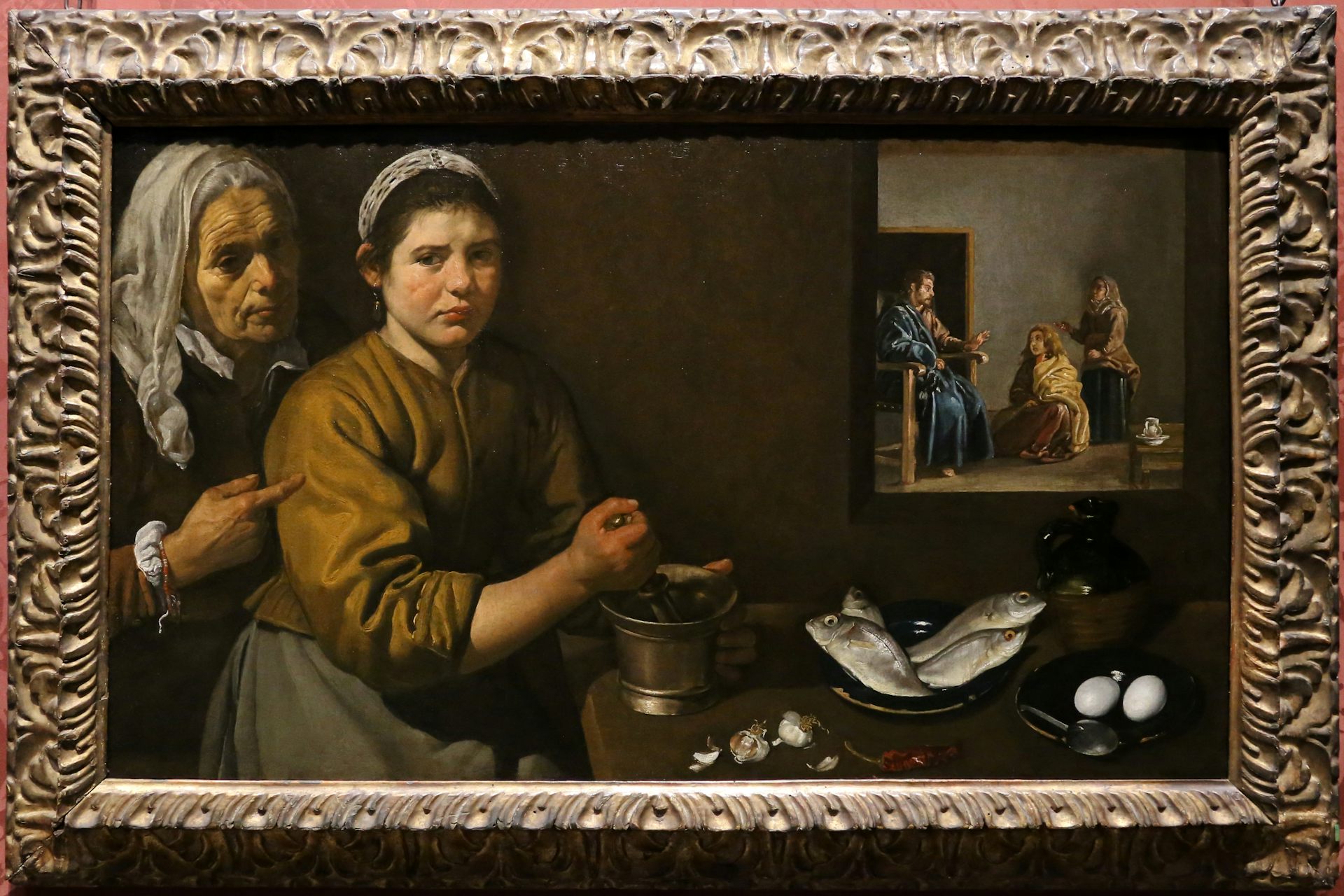 Painting of an older person pointing at a maid grinding garlic; a painting of Christ and Mary hangs above the plates of fish and eggs on the table