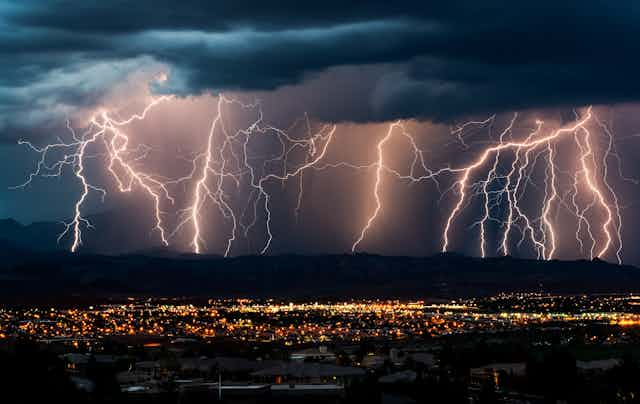 Multiple lightning strikes hit the mountains in the background beyond the lights of a town.