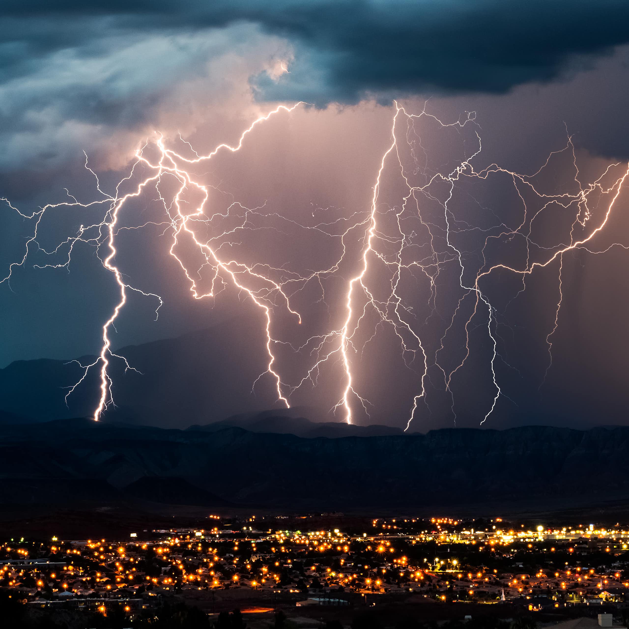 Multiple lightning strikes hit the mountains in the background beyond the lights of a town.