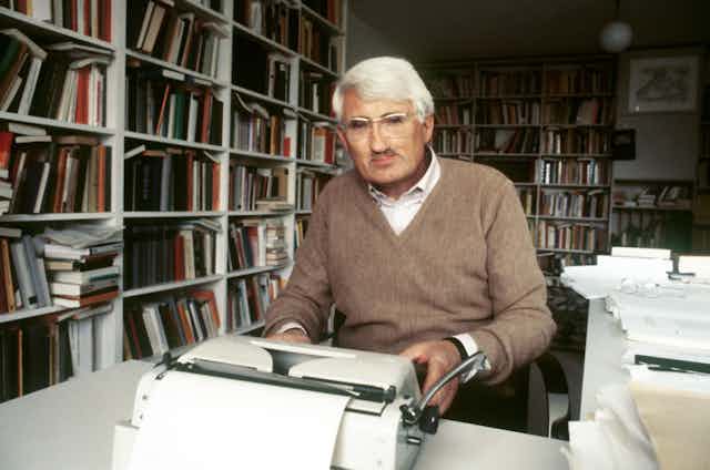 Man at his typewriter in a book-lined room.